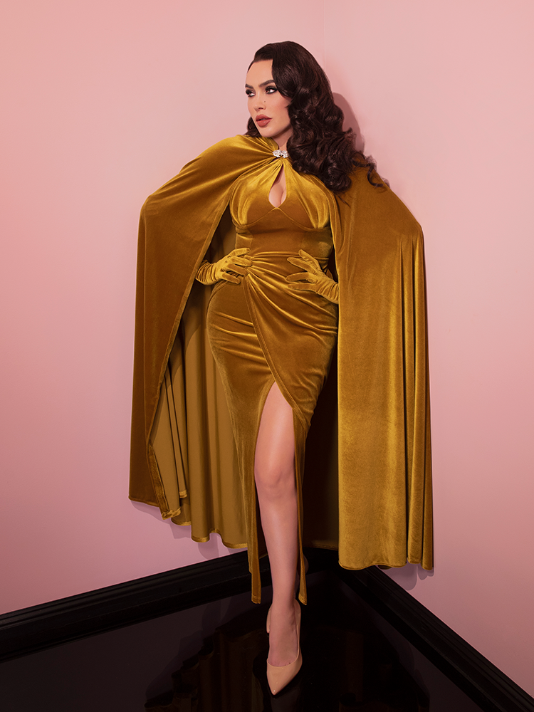 Micheline is in a pink room with her hands on her hips while wearing the Golden Era gown and glove set and matching cape.