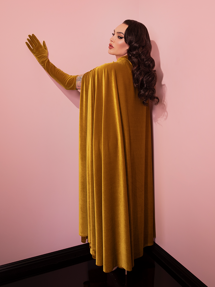 Micheline Pitt turned away from the camera to show off the back of the Golden Era Cape in Gold Velvet from retro clothing brand Vixen Clothing.