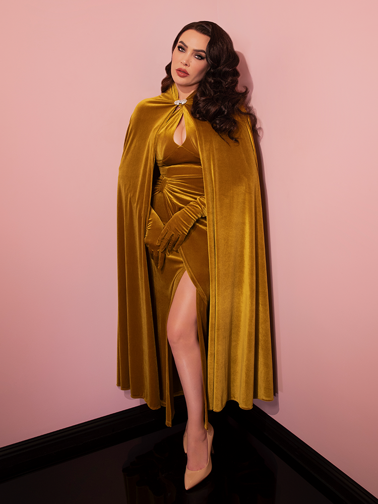 Micheline stands in the corner of a pink showroom wearing a golden velvet retro dress paired with a Golden Era Cape in Gold Velvet.