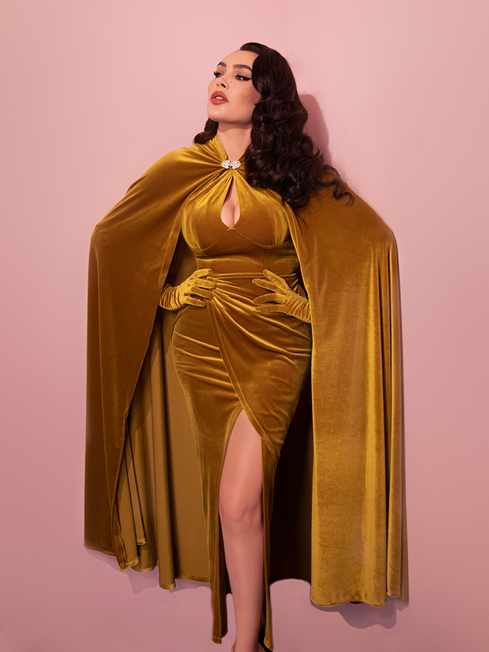 Micheline Pitt posing with her hands on her waist wearing the Golden Era Cape in Gold Velvet with matching retro dress.
