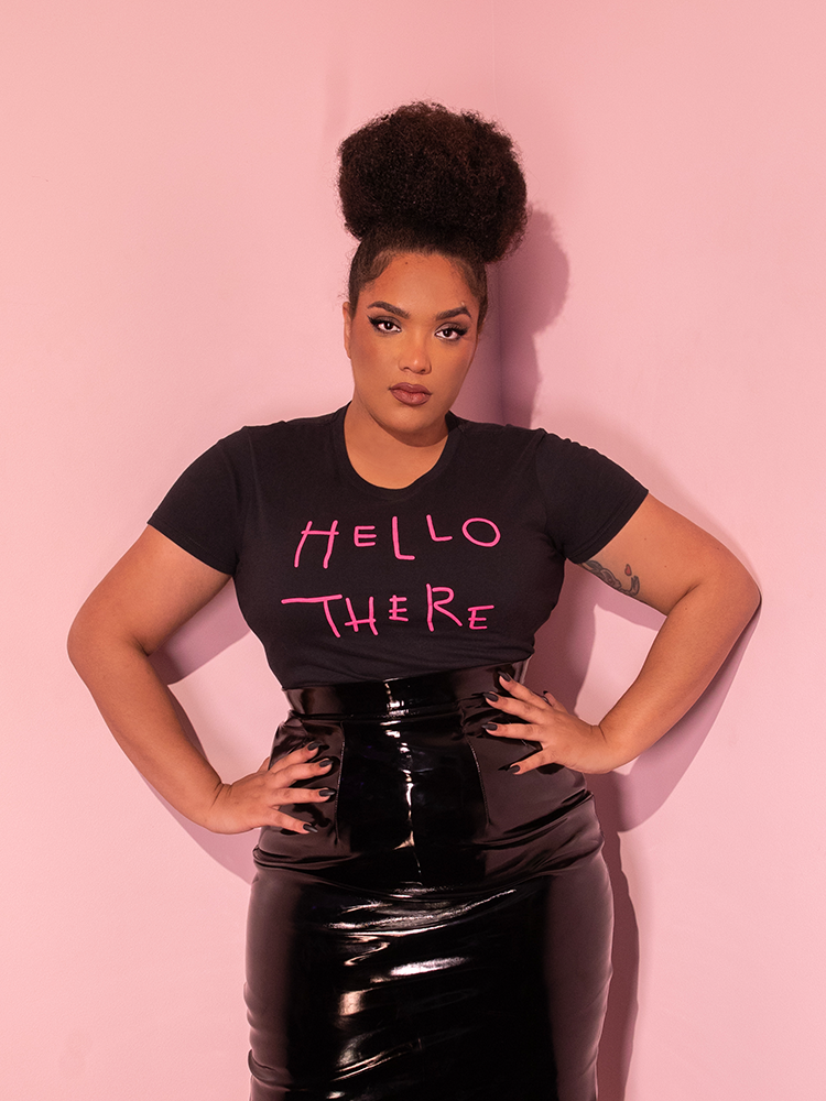 Model poses with her hands resting on her hips while wearing the BATMAN RETURNS™ Catwoman "Hello There" Women's Tee tucked into a black vinyl skirt.