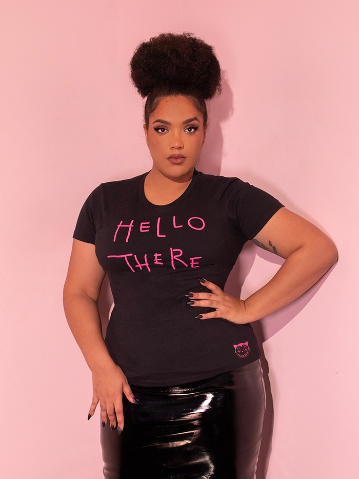 Female model posing with one hand on her hip and the other resting at the top of her thigh, wears the BATMAN RETURNS™ Catwoman "Hello There" Women's Tee over a black vinyl skirt.
