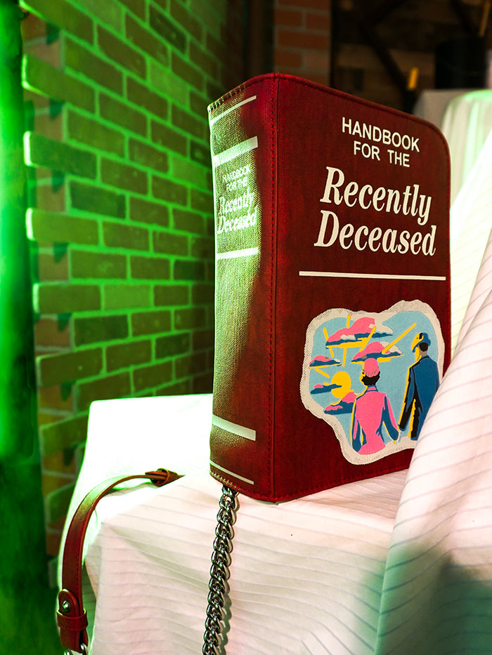 Product shot of the BEETLEJUICE™ Recently Deceased Book Bag in front of a green-lit brick wall.
