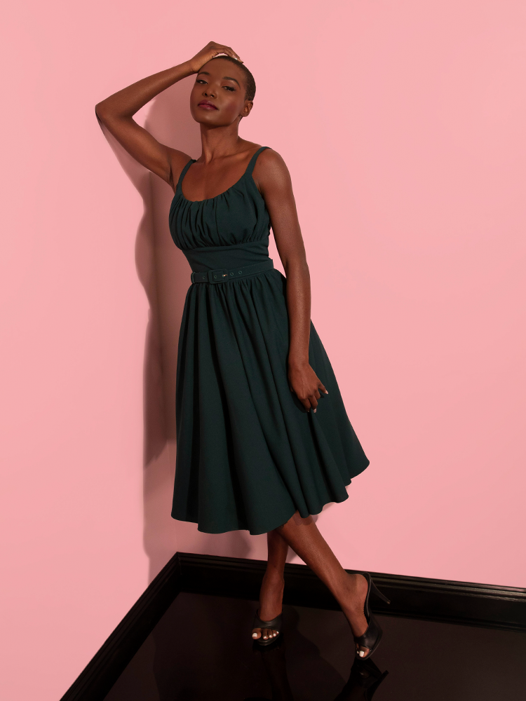 Model leans against the wall while wearing the Ingenue Dress in Hunter Green with black open toed shoes.