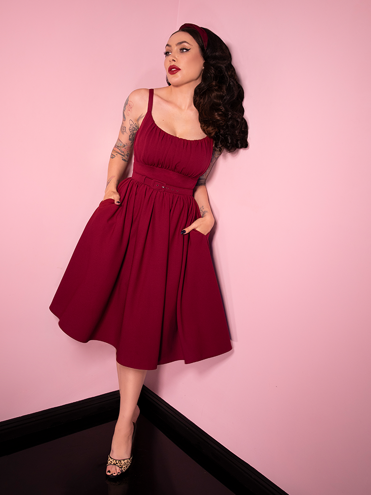 Full body shot of Micheline Pitt wearing a maroon colored retro style dress from her brand Vixen Clothing.