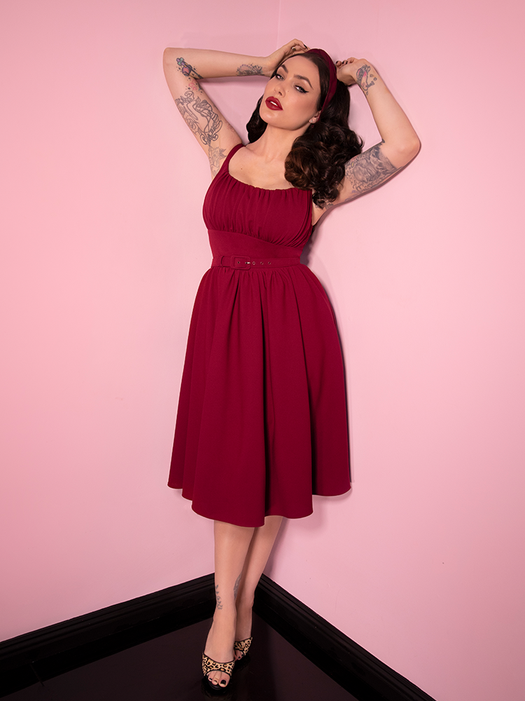 With her arms raised up, Micheline Pitt models the Ingenue Dress in Garnet from Vixen Clothing.