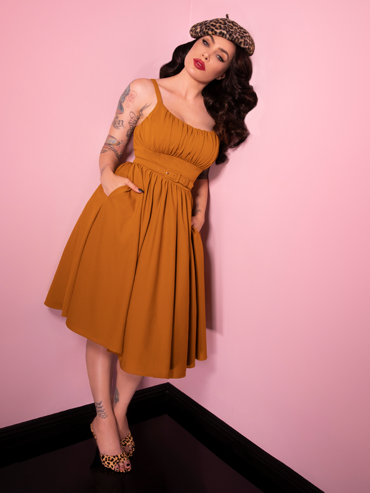 Leaning against the wall with her left shoulder, Micheline Pitt tucks her hands into her pockets of the Ingenue Dress in Vintage Mustard Print.