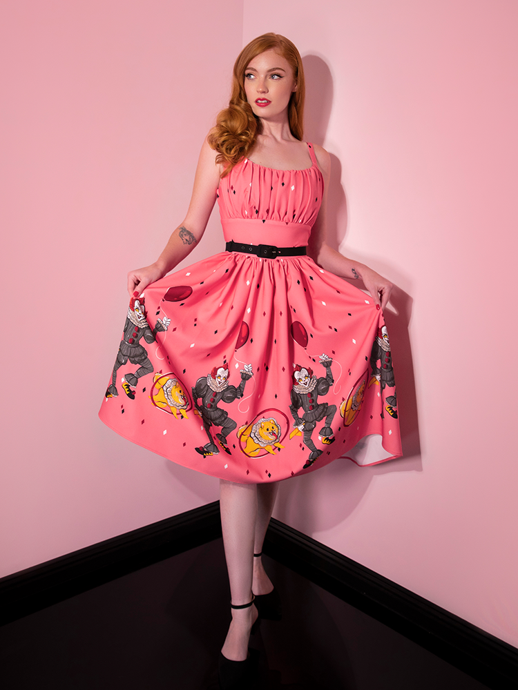 Looking to the side while she holds out her skirt, Emily models the Dancing Clown Ingenue swing dress in pink by Vixen Clothing.
