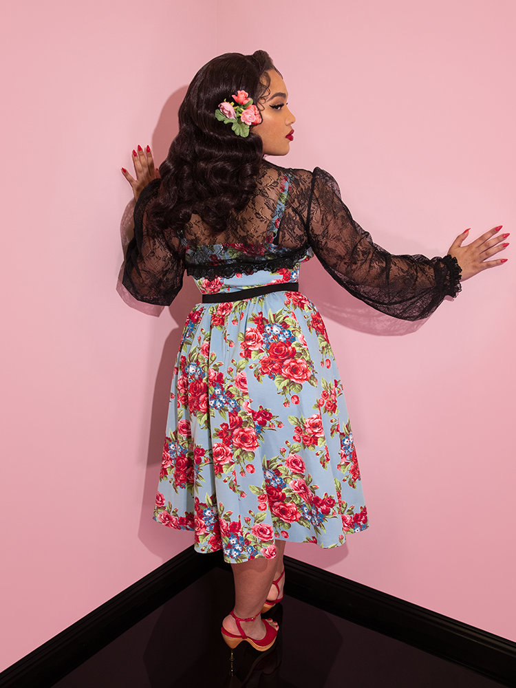 A back view of Ashleeta with flowers in her hair modeling the Vixen Clothing Ingenue dress in blue and red rose print paired with a black lace bolero.