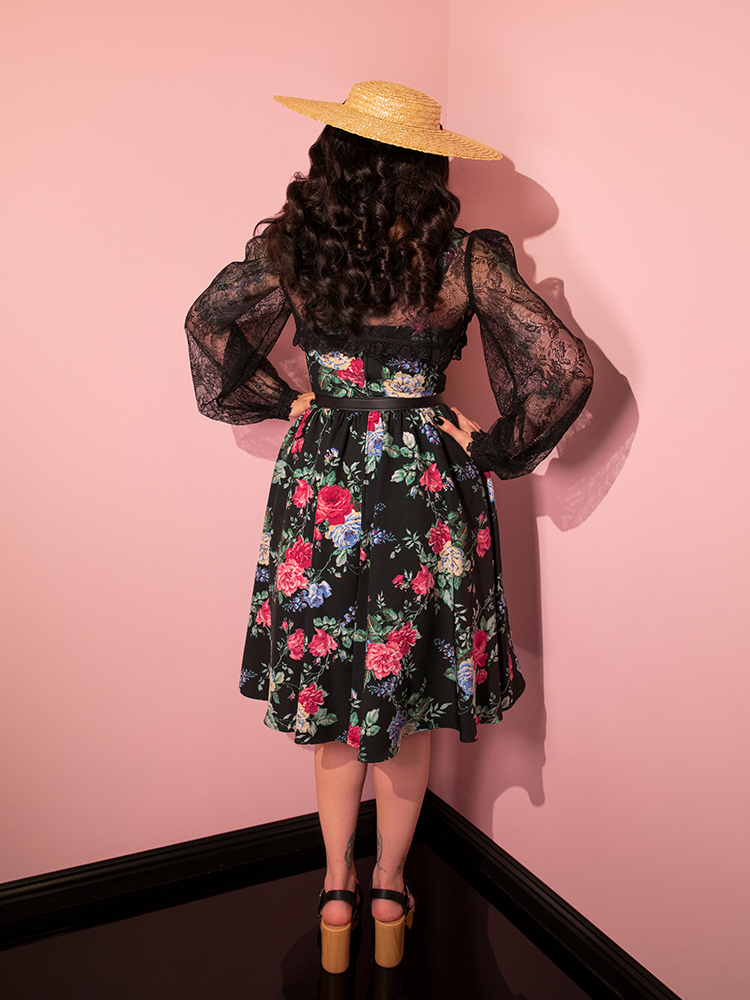 Micheline Pitt facing away from the camera to show off the back of the Ingenue Dress in Black Rose Print from retro dress company Vixen Clothing.