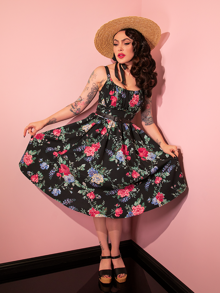 Micheline pulls the sides of her skirt taut to show off the print on the Ingenue Dress in Black Rose Print.