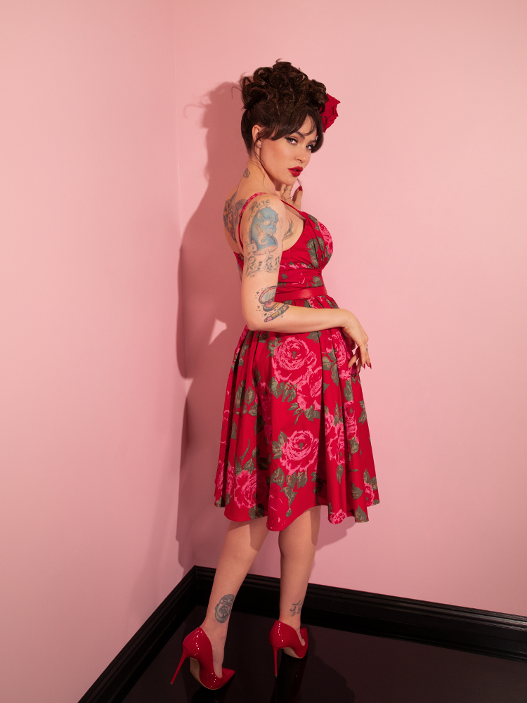 Micheline Pitt turned away from the camera looks back over her shoulder to show off the back of the Ingenue Swing Dress in Vintage Red Rose Print from Vixen Clothing.