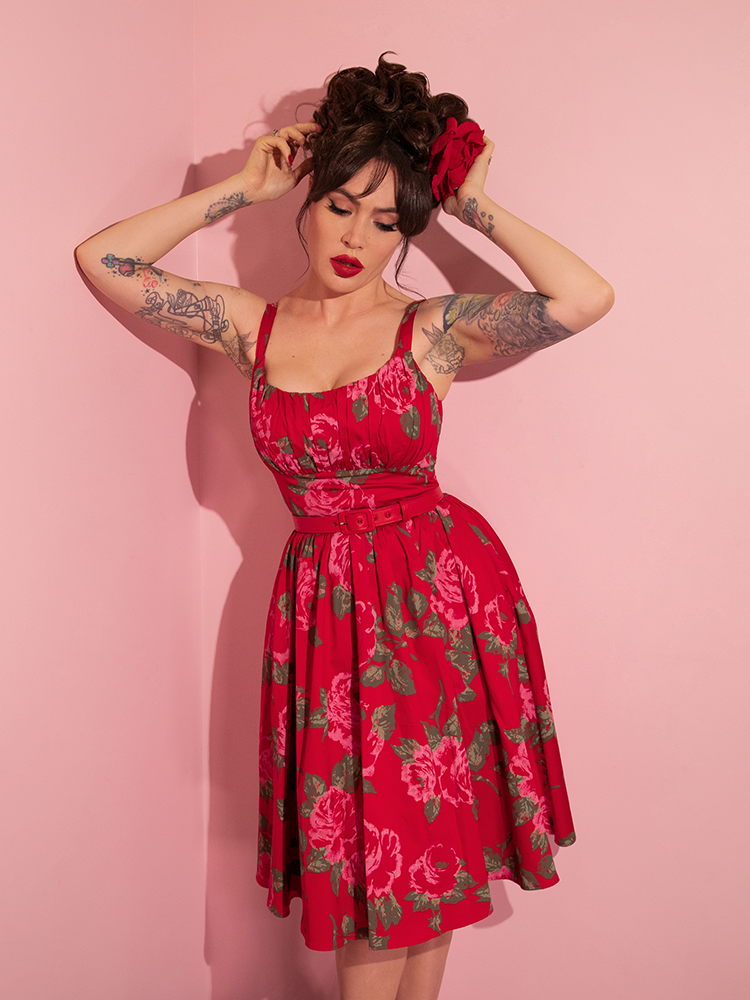 Micheline Pitt adjusts her hair while wearing the Ingenue Swing Dress in Vintage Red Rose Print from retro dress company Vixen Clothing.