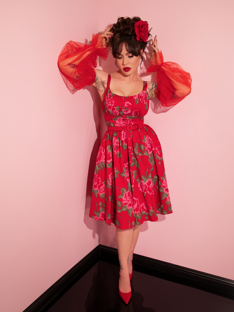 Full length shot of Micheline Pitt standing in the corner of a pink room wearing the Ingenue Swing Dress in Vintage Red Rose Print with red tulle sleeves.