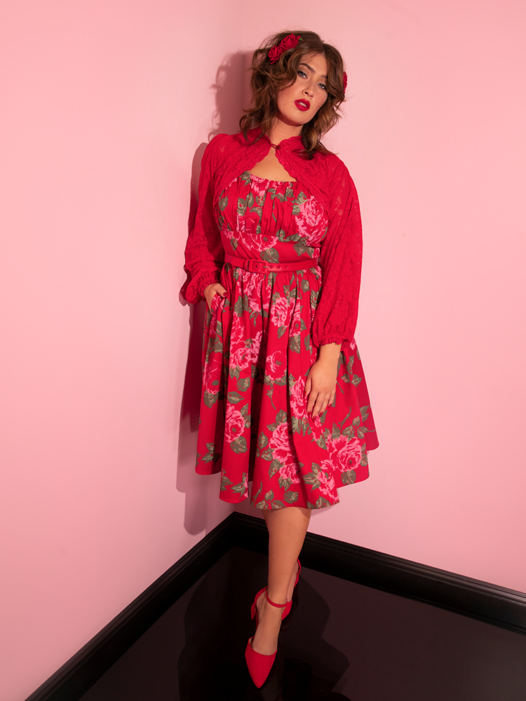 Francesca cooly walks with one hand in the pocket of her Ingenue Swing Dress in Vintage Red Rose Print from retro dress company Vixen Clothing.