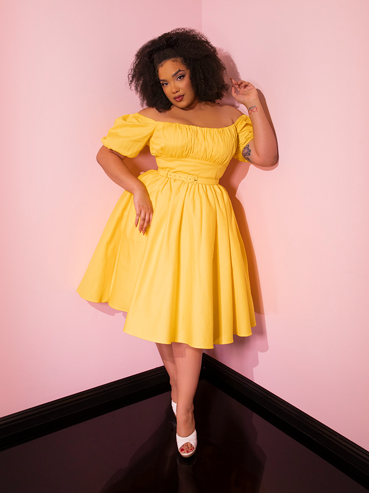 Full length shot of model posing in the corner of a room in the Lakeland Dress in Yellow from Vixen Clothing.