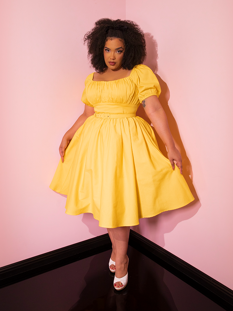 Full length shot of model gently pulling out the sides of the Lakeland Dress in Yellow from Vixen Clothing.