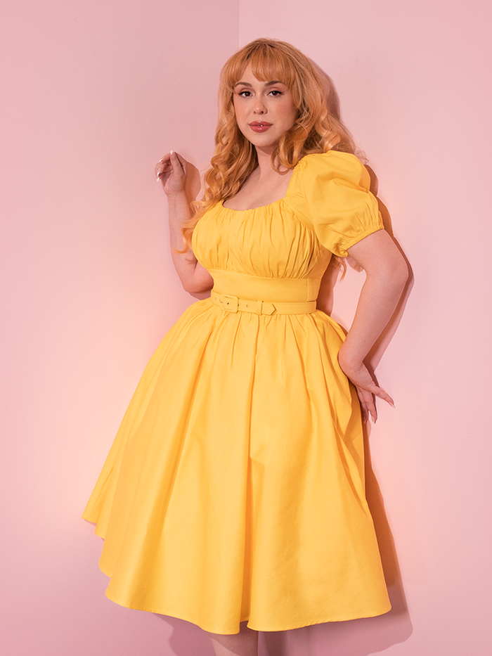 Auburn haired model wearing the Lakeland Dress in Yellow while leaning up against a wall.