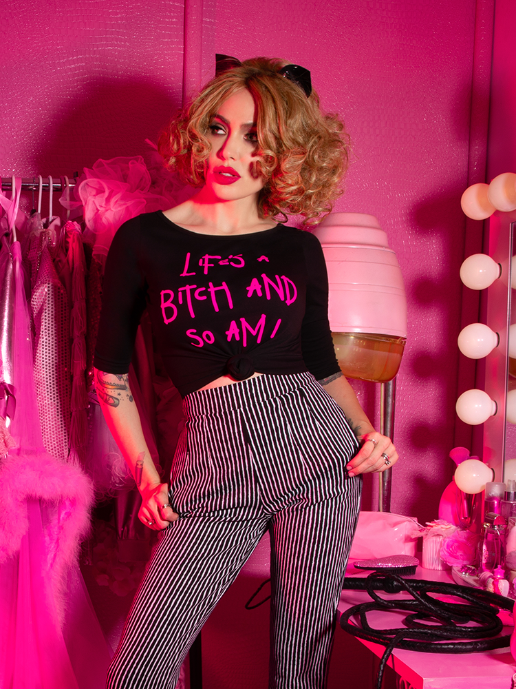 Micheline Pitt standing in a pink dressing room wearing black and white striped pants and the Miss Kitty Life's a Bitch Raglan T-Shirt.