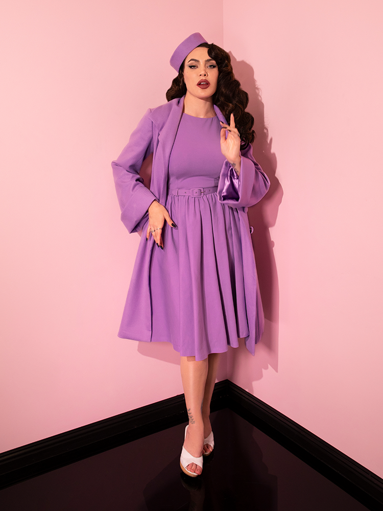 Micheline Pitt posing seductively while wearing the Avon Swing Dress in Lilac from retro dress company Vixen Clothing.