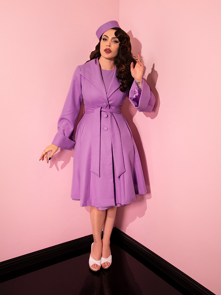 Micheline Pitt posing with one hand raised up to her head and the other posed by her hip, wears the The Lili Coat in Lilac.