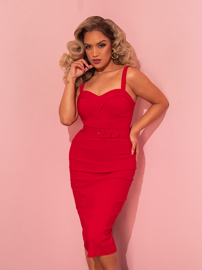 A stunning female model adds a playful touch to her allure while posing in the Maneater Wiggle Dress in Red from the retro clothing brand Vixen Clothing.