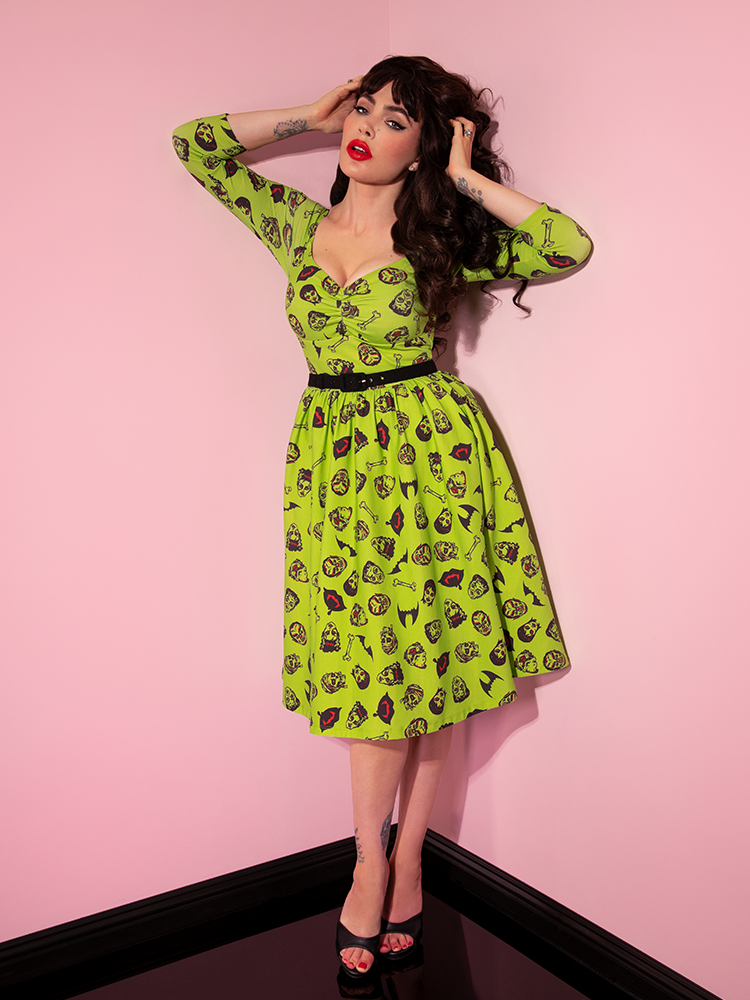 Micheline Pitt posing with her arms stretched up and fingers in her hair, wearing the Wicked Swing Dress in Vintage Monster Mash Print. 