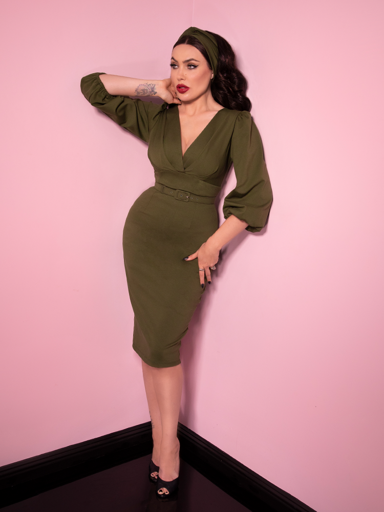 Micheline Pitt looking away from camera modeling the Bawdy wiggle dress in olive green paired with a matching headband.