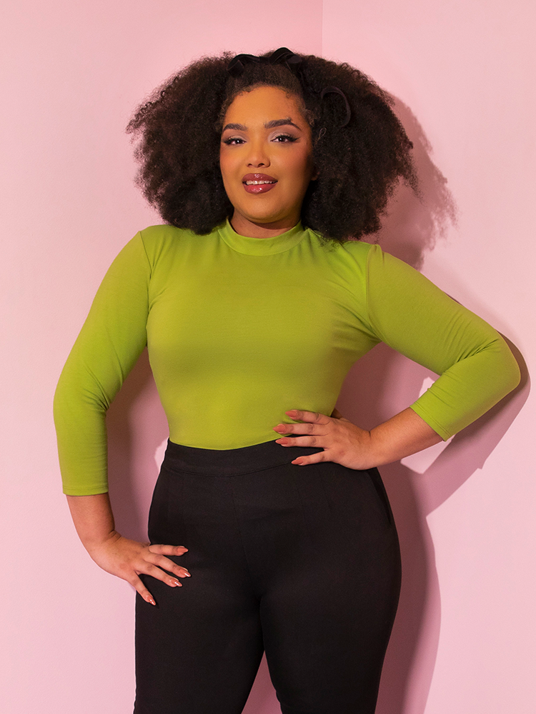 Model smiles as she places her hands on her hips while wearing the  Bad Girl 3/4 Sleeve Top in Avocado Green tucked into black cigarette pants.