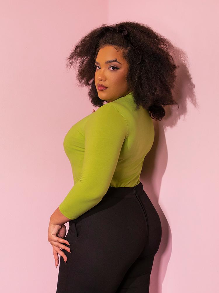 Model turns to the side, while look into the camera, gives the viewer a profile view of the  Bad Girl 3/4 Sleeve Top in Avocado Green she's wearing while tucked into pants. 