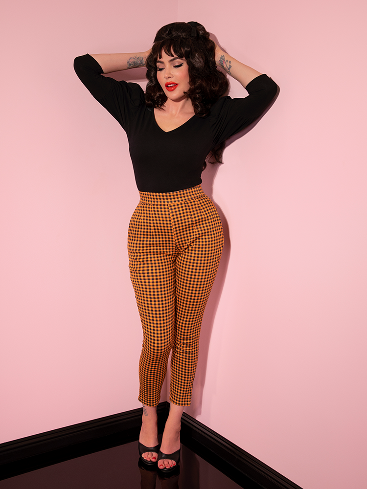 Micheline Pitt holding her hands behind her head poses in the Cigarette Pants in Orange Pumpkin Gingham.