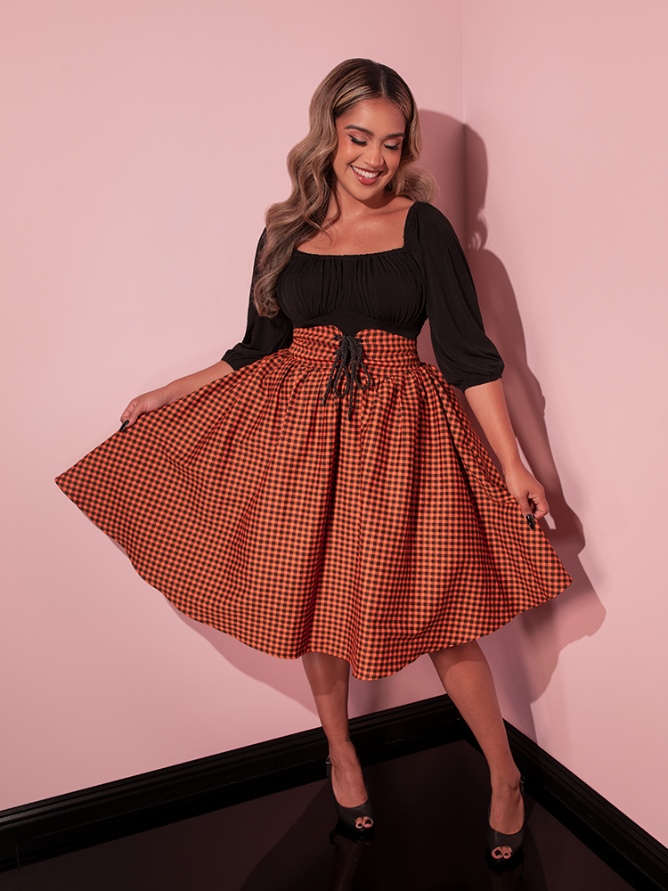 Female model, smiling and looking down, holds out the sides of the Corset Skirt in Orange Pumpkin Gingham.