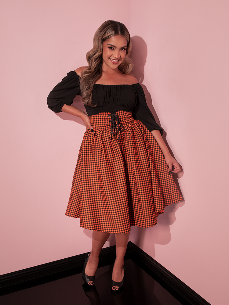 Model smiling and looking at the camera while wearing the Corset Skirt in Orange Pumpkin Gingham with a black long sleeve top tucked into the skirt.