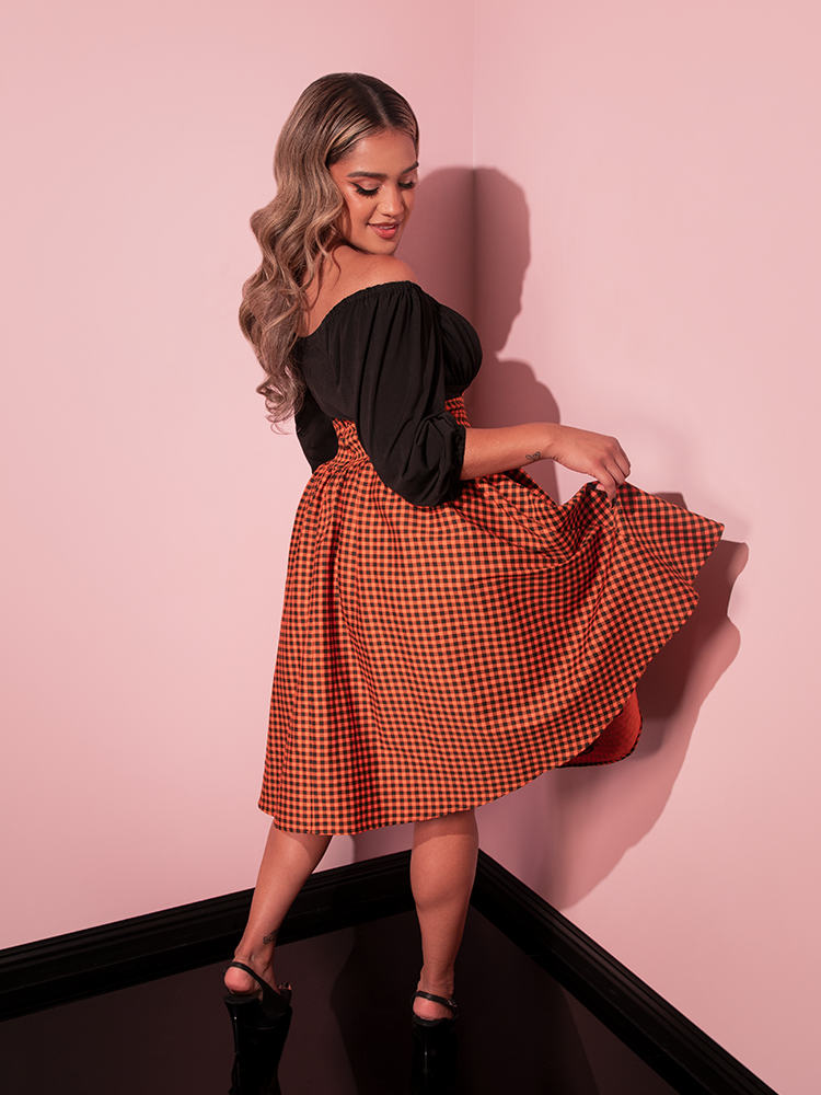 Model turned away from the camera, but looking over her shoulder at the back of the Corset Skirt in Orange Pumpkin Gingham from retro clothing brand Vixen Clothing.