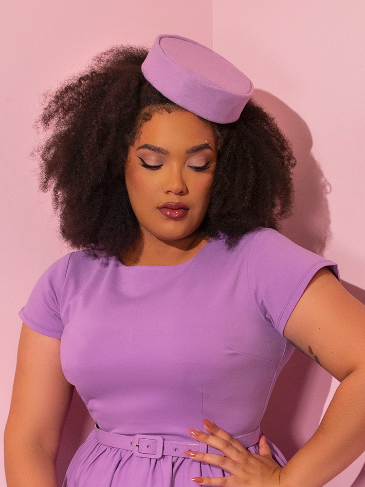 Waist level shot of model wearing a lilac colored retro style dress with matching Vintage Pillbox Hat in Lilac.