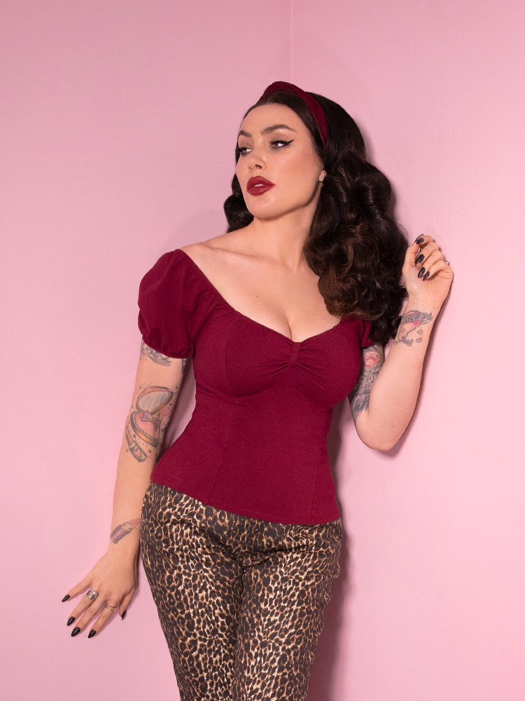 Micheline Pitt posing against pink walls while wearing a vintage inspired outfit from Vixen Clothing featuring their Powder Puff Top in Burgundy.