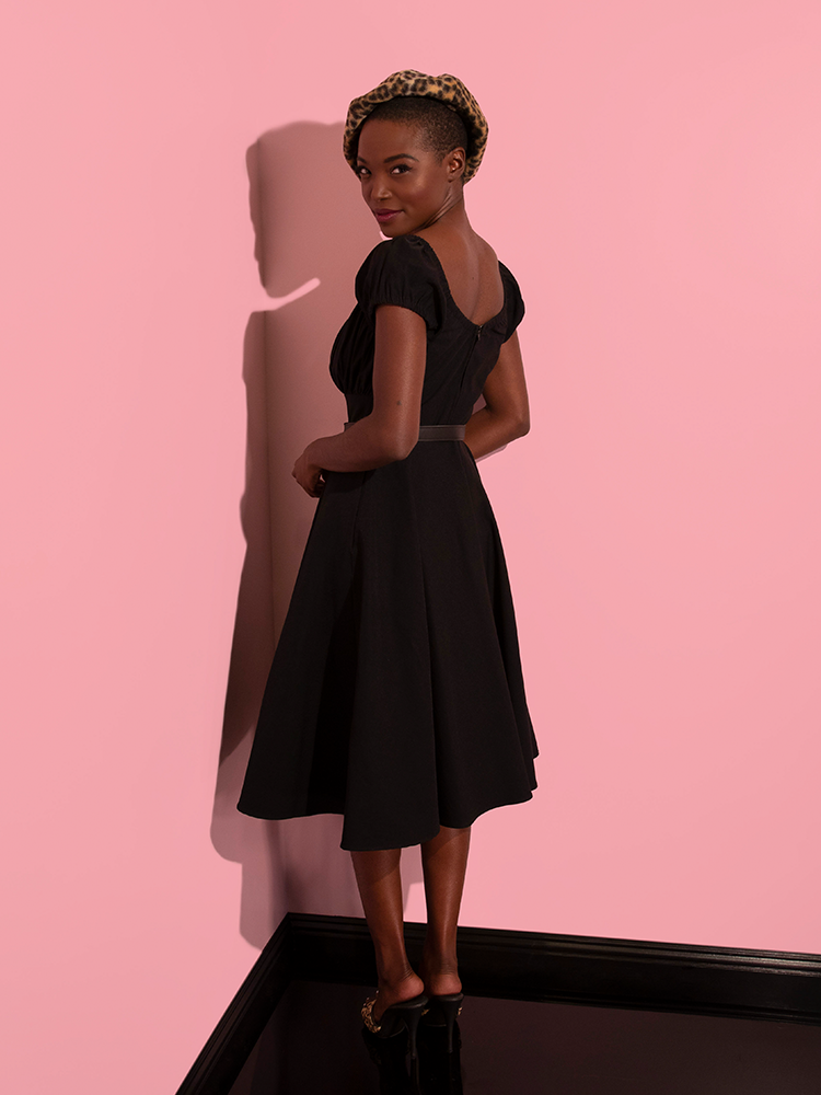 Turned away from the camera but looking back, an African-American model wears the Peasant Swing Dress in Black from retro clothing brand Vixen Clothing.