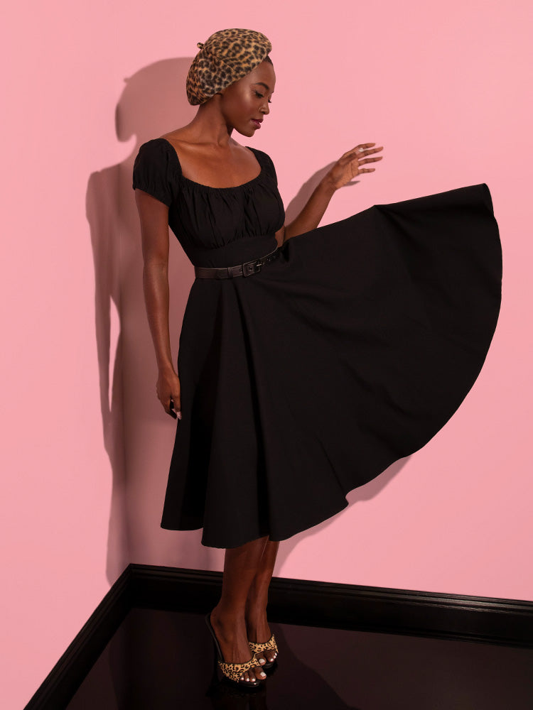 Full length image of model posing in the Peasant Swing Dress in Black while the skirt section of the dress is tossed outward.