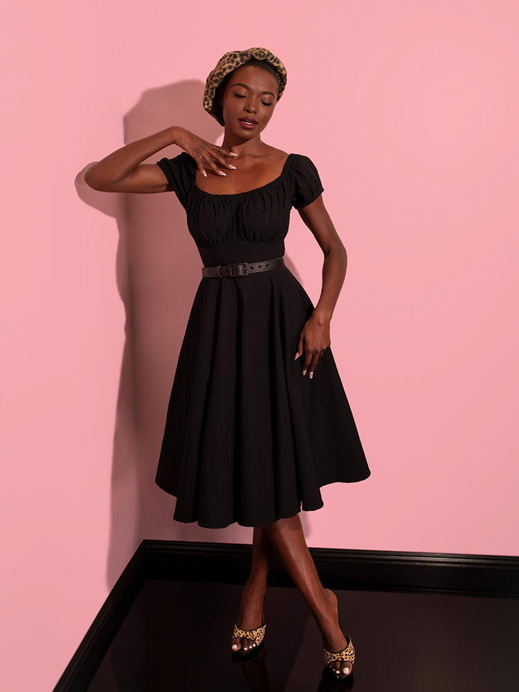 Model posing with one hand on her placed delicately on her chest and the other resting at her side while wearing the Peasant Swing Dress in Black. 