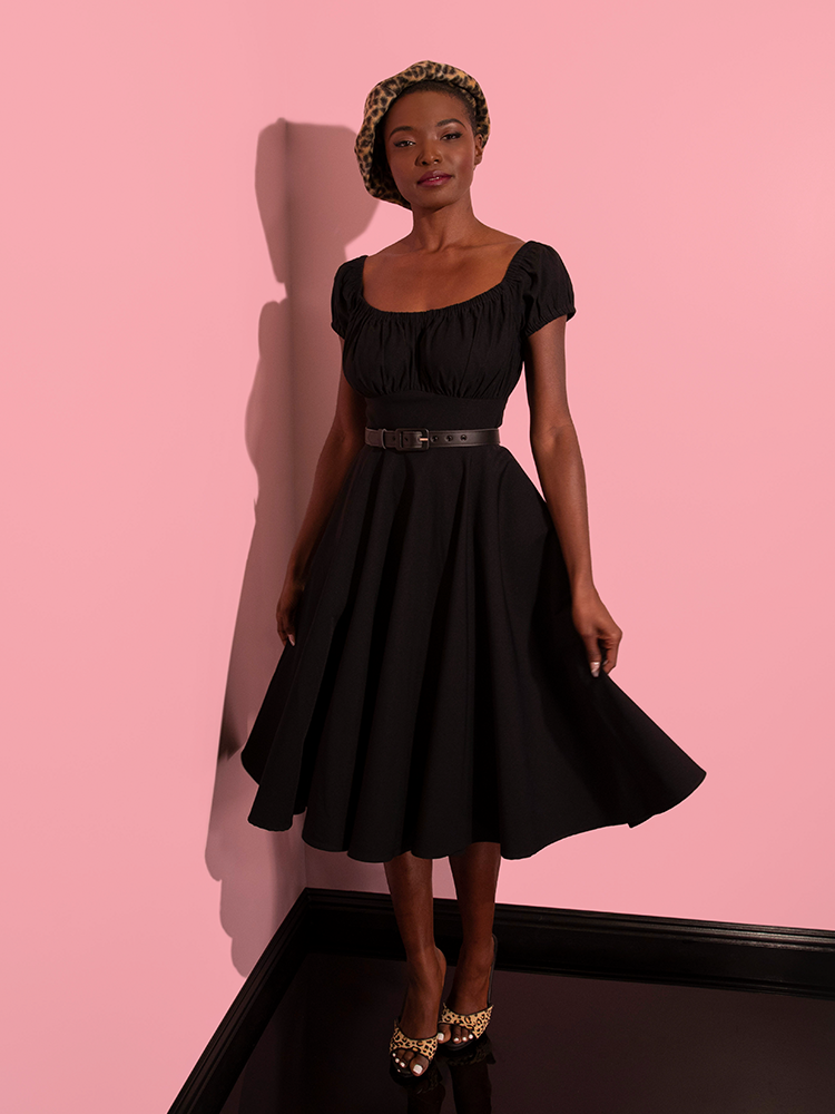 Model, smiling slightly, looks on to the camera while wearing the Peasant Swing Dress in Black. 