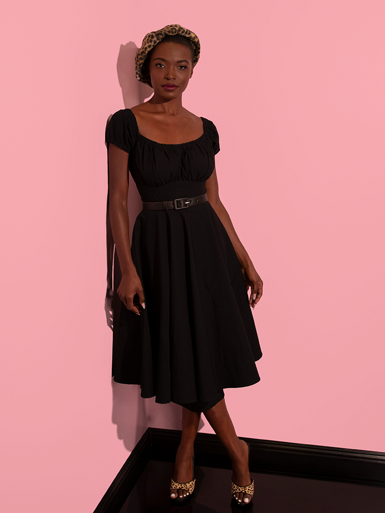 African American model is photographed looking at the camera while wearing the Peasant Swing Dress in Black paired with a leopard print beret. All items are available from Vixen Clothing.