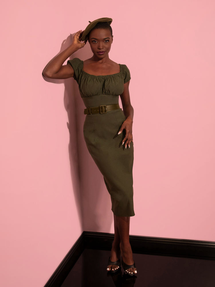 Full length shot of model wearing the Peasant Wiggle Dress in Olive Green with removable faux leather belt and matching color beret. All items from retro clothing company Vixen Clothing.