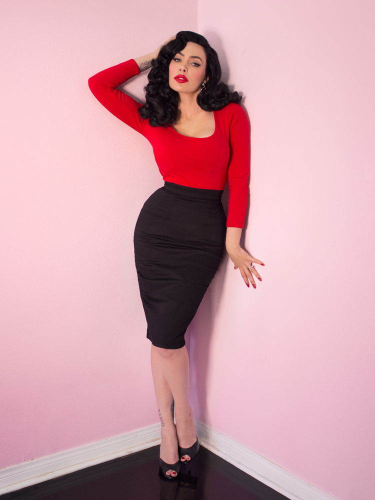 A full length photo of Micheline Pitt modeling the Troublemaker top in red by Vixen Clothing paired with a black pencil skirt.