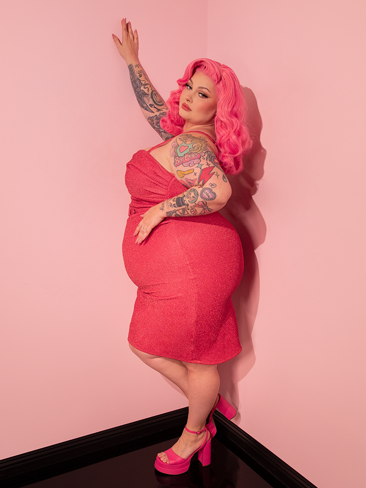 The Candy Pink Lurex Jawbreaker Wiggle Dress, a bewitching piece from Vixen Clothing's retro clothing line, accentuates the female model's sexy pose, leaving a lasting impression on the audience.
