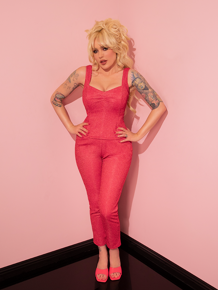 The retro style model exudes confidence and charm as she proudly presents the Cigarette Pants in Candy Pink Lurex by Vixen Clothing, a renowned vintage clothing maker.