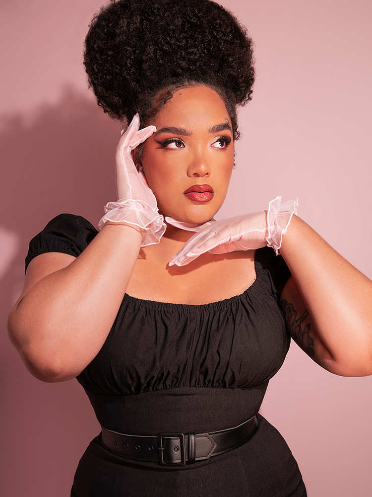 The Sheer Wristlet Gloves in Pink worn by Vixen Clothing model, Ashleeta, while also wearing a low-cut black top and black pants.