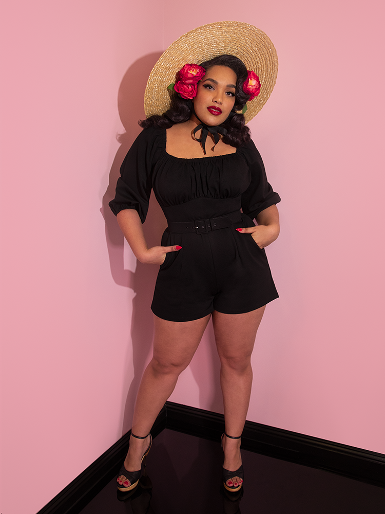 Full length image of Ashleeta in a retro inspired outfit featuring black playsuit and natural colored sunhat.