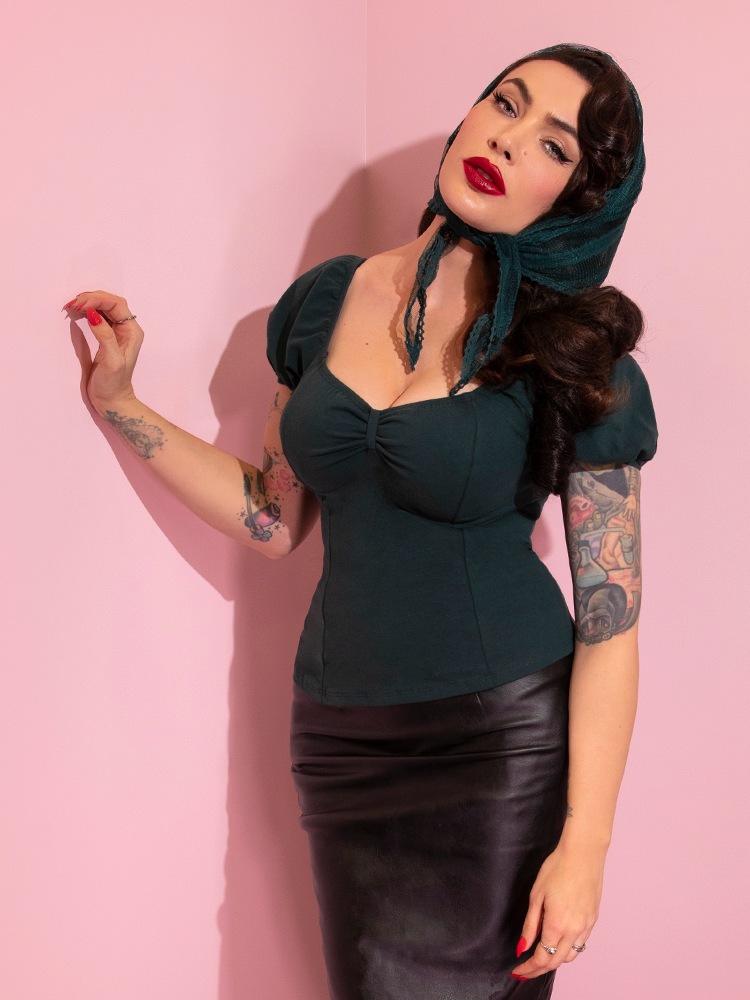 A closeup of Micheline Pitt looking at the camera modeling the Powder Puff top in hunter green untucked paired with a black pencil skirt and green head scarf.