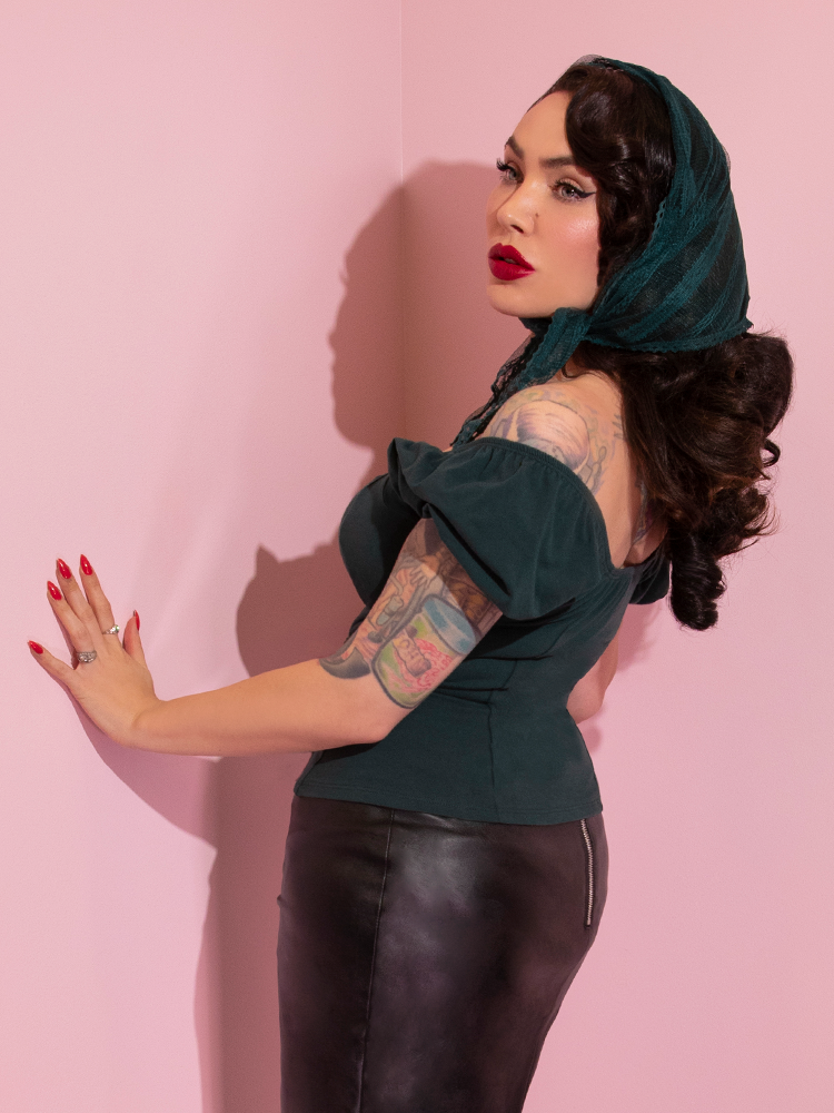 A side view of Micheline Pitt looking away from camera modeling the Powder Puff top in hunter green paired with a black pencil skirt and green head scarf.