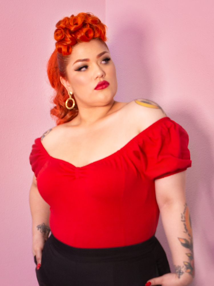 Evelyn looking off camera with her hands in her pockets modeling the Powder Puff top in red by Vixen Clothing.